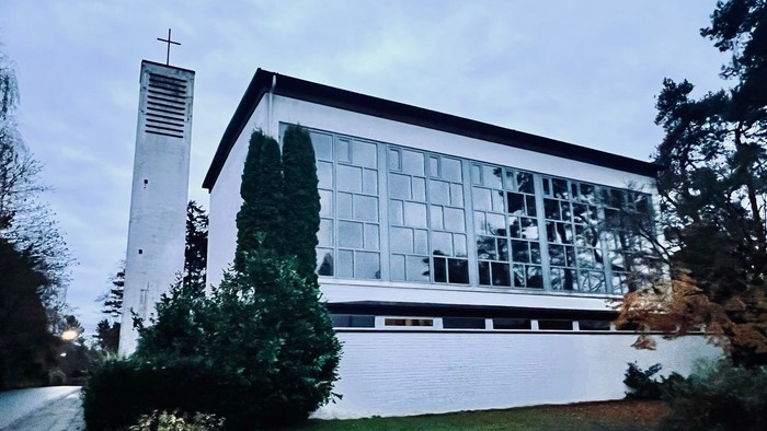 Moderne Kirchengebäude mit großen Glasfenstern und einem hohen Glockenturm neben üppigem Grün.
