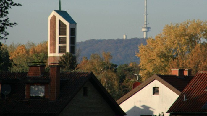 Städtische Skyline mit Kirchturm, Wohnhaustdächern und einem hohen Sendemast vor herbstlichem Laub.