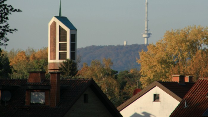 Städtische Skyline mit Kirchturm, Wohnhaustdächern und einem hohen Sendemast vor herbstlichem Laub.