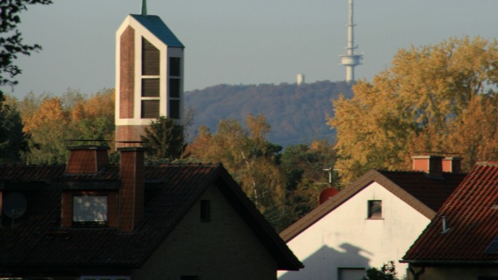 Städtische Skyline mit Kirchturm, Wohnhaustdächern und einem hohen Sendemast vor herbstlichem Laub.