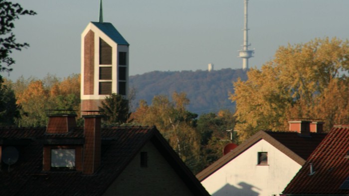 Städtische Skyline mit Kirchturm, Wohnhaustdächern und einem hohen Sendemast vor herbstlichem Laub.