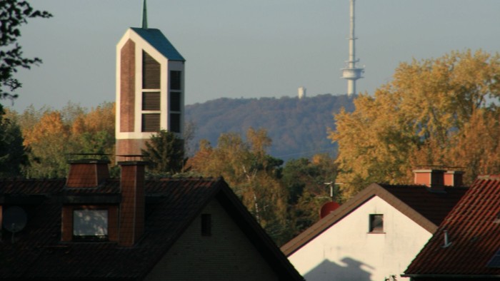 Städtische Skyline mit Kirchturm, Wohnhaustdächern und einem hohen Sendemast vor herbstlichem Laub.