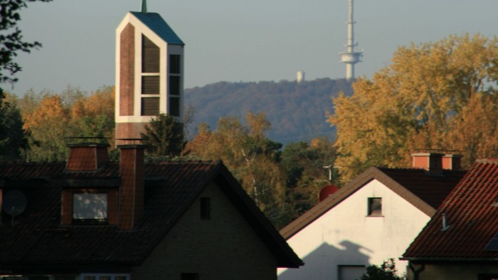 Städtische Skyline mit Kirchturm, Wohnhaustdächern und einem hohen Sendemast vor herbstlichem Laub.