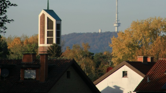 Städtische Skyline mit Kirchturm, Wohnhaustdächern und einem hohen Sendemast vor herbstlichem Laub.