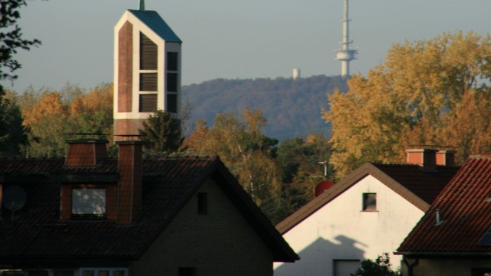 Städtische Skyline mit Kirchturm, Wohnhaustdächern und einem hohen Sendemast vor herbstlichem Laub.