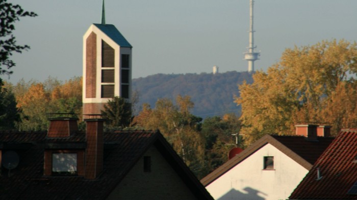Städtische Skyline mit Kirchturm, Wohnhaustdächern und einem hohen Sendemast vor herbstlichem Laub.