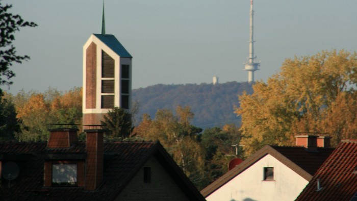 Städtische Skyline mit Kirchturm, Wohnhaustdächern und einem hohen Sendemast vor herbstlichem Laub.