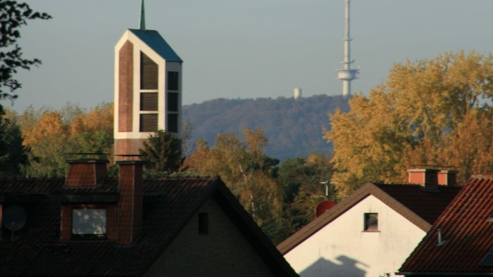 Städtische Skyline mit Kirchturm, Wohnhaustdächern und einem hohen Sendemast vor herbstlichem Laub.