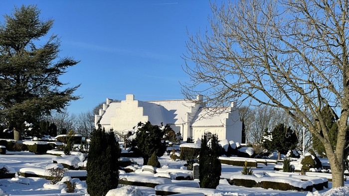 Et fredfyldt vintermotiv med et hvidt hus omgivet af sneklædte buske og nøgne træer under en klar blå himmel.