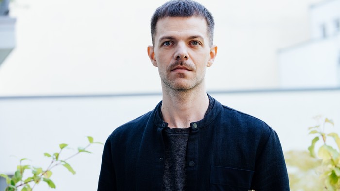 Ein Mann in dunkler Kleidung steht vor einem unscharfen Hintergrund aus Gebäuden und Grünpflanzen.