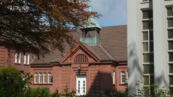 Ein rotes Backsteingebäude mit grünem Turm und weißen Fenstern, umgeben von Bäumen und Büschen.