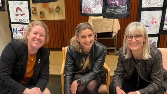 Tre smilende kvinder sidder sammen ved et bord med papirer, glas og kunstværker i baggrunden.