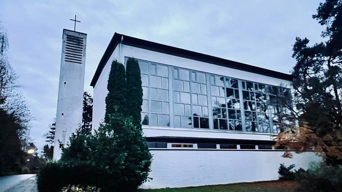 Moderne Kirchengebäude mit großen Glasfenstern und einem hohen Glockenturm neben üppigem Grün.