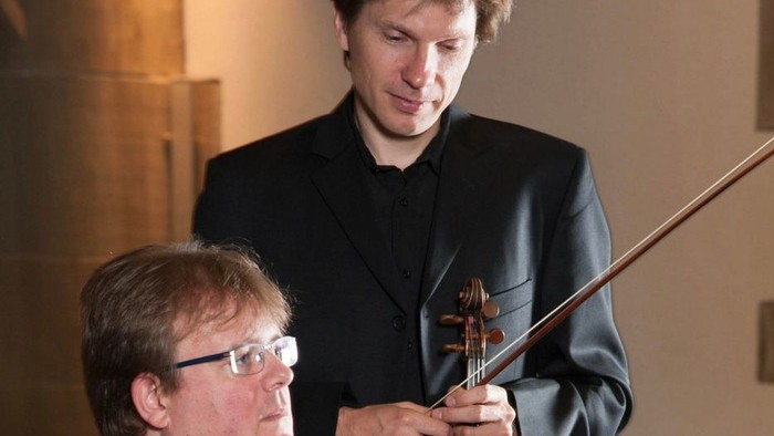 Zwei Musiker, einer mit einem Cello, wirken konzentriert während einer Aufführung oder Probe.