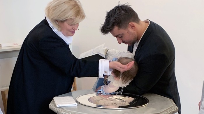 En præst og en kvinde holdes et barn over en fontæne under en religiøs ritual.