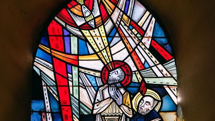 Buntglasfenster, das eine Heiligengestalt mit Heiligenschein zeigt, die eine andere Person segnet, in leuchtenden, kunstvollen Farben und geometrischen Mustern.