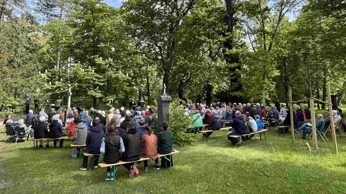 Eine große Menschenansammlung im Freien, die auf Bänken in einer üppigen, grünen Parklandschaft sitzt.