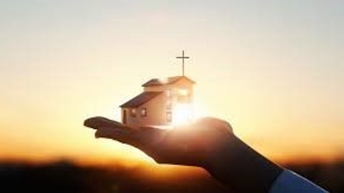 Eine Hand hält ein kleines Haus mit Kreuz vor einem Sonnenuntergang.