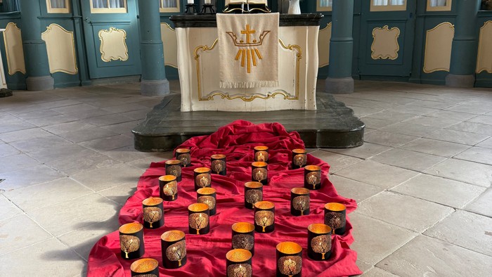 Elegante Altaraufstellung mit zahlreichen goldenen Votivkerzen auf einem roten Tuch, eingerahmt von einem dekorativen Kircheninnenraum.