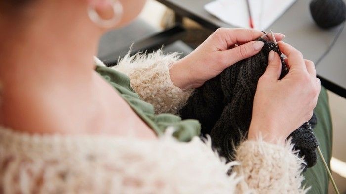 En person sitter og stråler en sort trøje med nåle og garn.