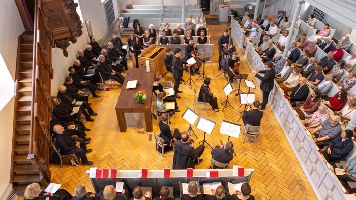 Großer Raum mit vielen Menschen, die auf Stühlen sitzen und Notenblätter halten, ein Orchester proben.