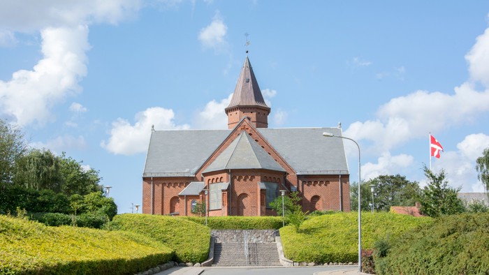 En rød kirke med spiret og en flagstang med danskirkeflag ved siden af.