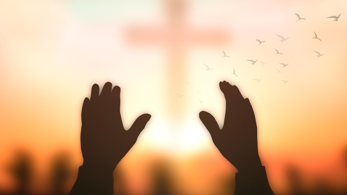 Hands raised towards a glowing cross at sunset with birds flying.
