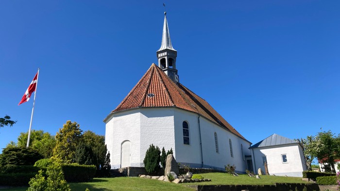 En hvid kirke med spiret og rødt tag, flagstang med danskirkeflag ved siden.