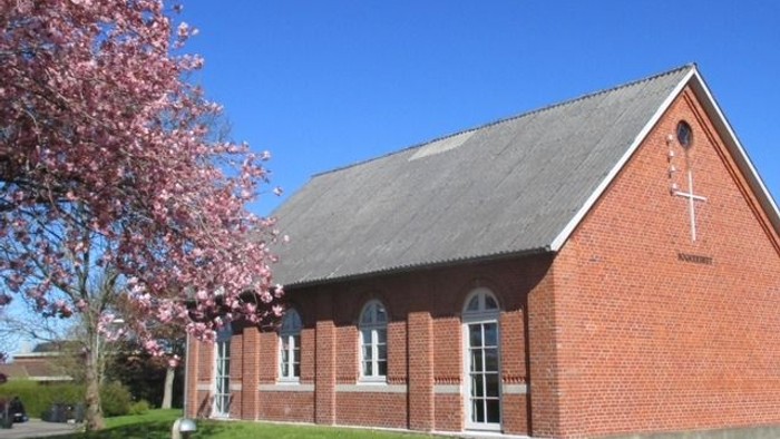 En rød murstenskirke med kryds og blomstrende træ foran