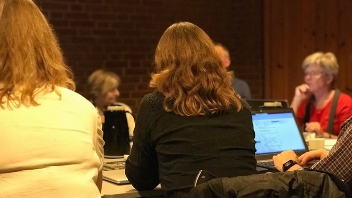 En gruppe af personer sidder ved bord i et rum med mur af røde mursten.