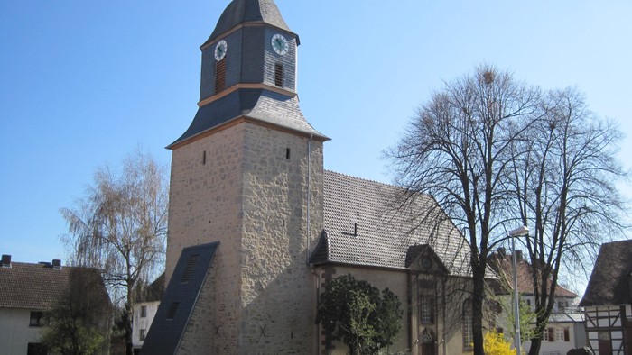 Außenansicht der evangelischen Kirche in Oberlistingen. Das Gebäude besteht aus hellem Naturstein mit einem hohen Turm, der eine schieferverkleidete, geschwungene Dachhaube mit Uhr trägt. Das Kirchenschiff hat ein dunkles Ziegeldach. Vor der Kirche stehen