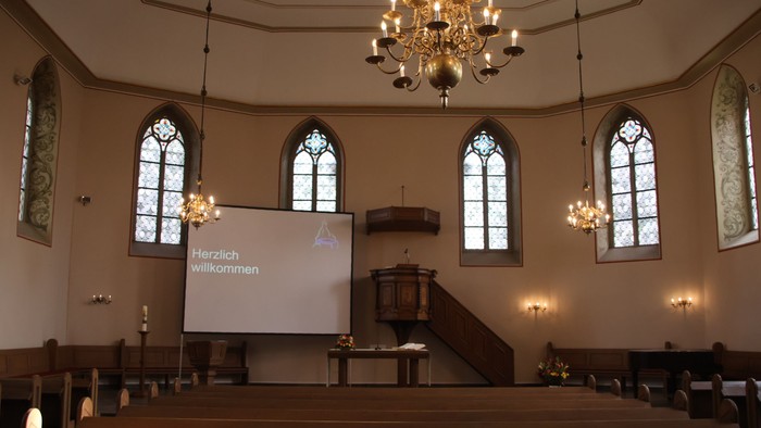 Kircheninneres mit Buntglasfenstern, Kronleuchtern und einem Bildschirm, der eine Willkommensbotschaft anzeigt.