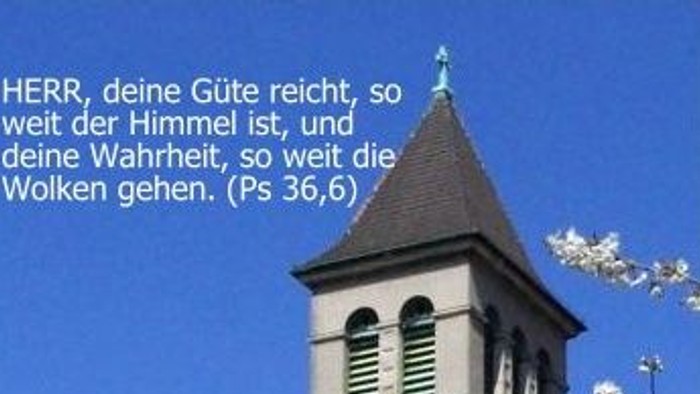 Ein Kirchturm mit blühenden Bäumen und einem biblischen Zitat auf Deutsch vor einem klaren blauen Himmel.