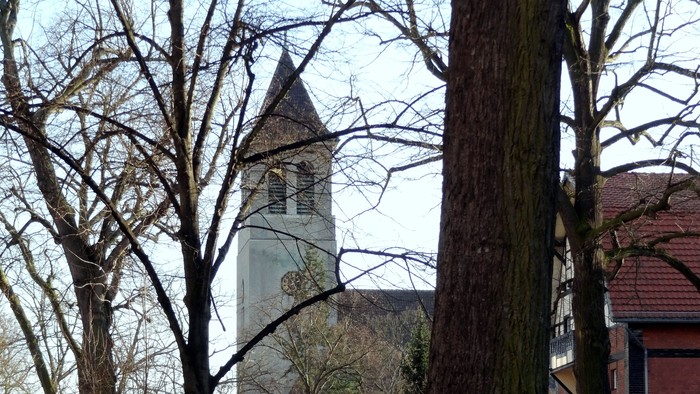 Ein Kirchturm ragt hinter Bäumen empor, daneben ein rotes Gebäude.