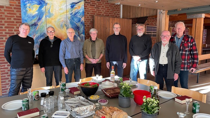 En gruppe mænd står sammen ved et bord med mad og drikkevarer i et rum med murmalet og store billeder.