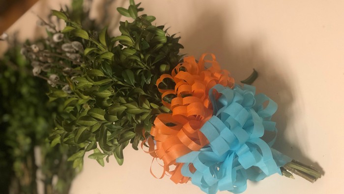 Dekorative Anordnung mit Grünpflanzen, orangefarbenen und blauen Lockenbändern, die mit einer Schleife gebunden sind.