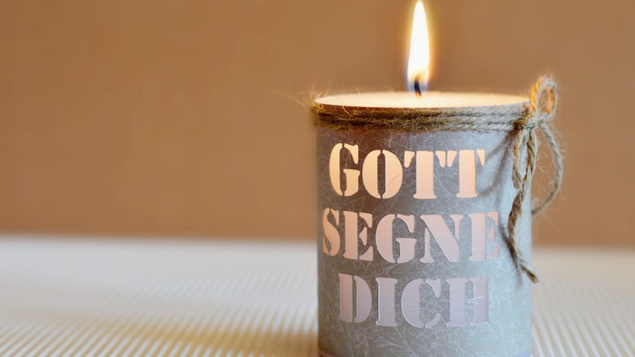 Kerze in Metallbehälter mit deutschem Text, angezündet und mit einer Schnur umwickelt.
