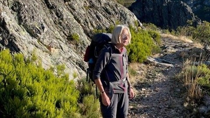 En vandringsmand med en rygsæk bevæger sig hen over en ujævn, solbeskinnet bjergsti omgivet af klippefyldt terræn og sparsom vegetation.