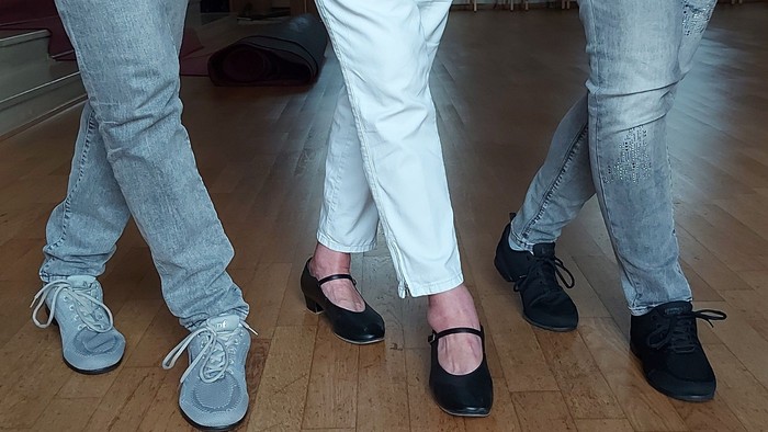 Drei Personen stehen auf einem Holzboden, ihre Schuhe sind zu sehen.