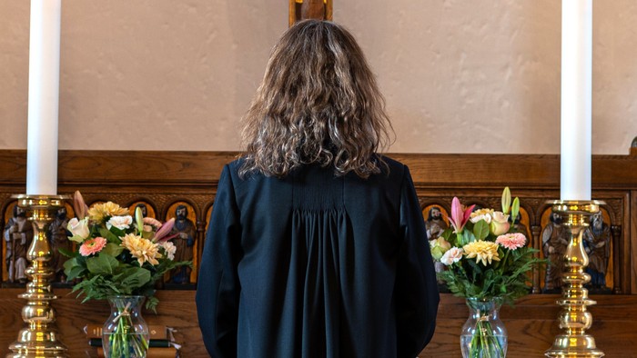 En person i mørkt kapittel ser op mod alteret i en kirke med lysede steariner og blomster.