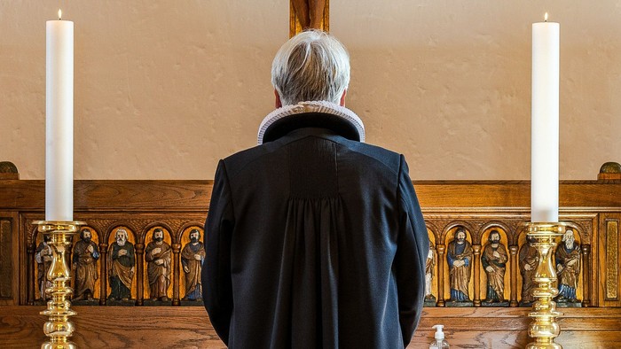 En ældre mand sitter alene i en kirke, med to lysende lysstake på hver side.