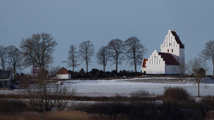 En hvid kirke med tårn og et mindre hus i en snedækket landskab med træer.