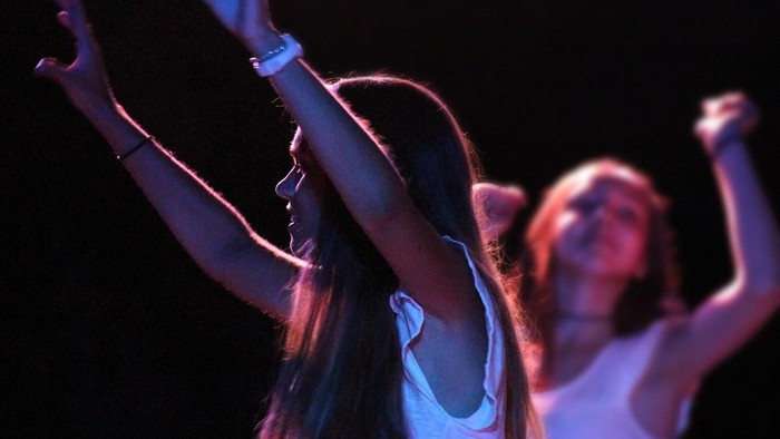 Zwei Frauen, die mit erhobenen Armen tanzen, eine trägt ein weißes Hemd.