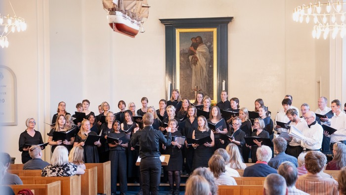 Et stort kor optræder i en kirke og synger efter noder, mens publikum lytter opmærksomt.