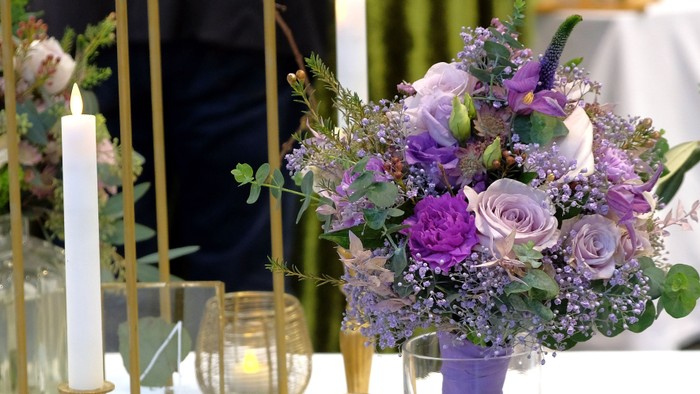 Im Vordergrund rechts ein Blumenstrauß in Lila, auf einem mit weißen Stabkerzen in goldenen Leuchtern geschmückten Hochzeitstisch