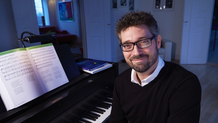 En mand med briller sidder ved et klaver med noder og smiler varmt i et hyggeligt rum.