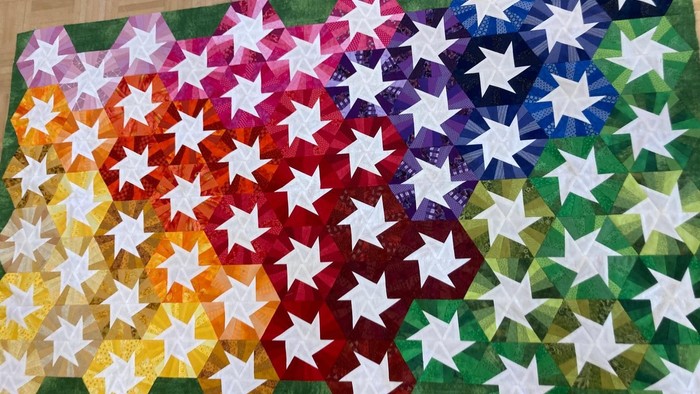 Eine Patchworkarbeit in Regenbogenfarben, viele Sechsecke von gelb über orange, rot, blau und grün, immer mit einem weißen Rad in der Mitte.