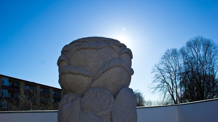 En stenstat af en figur med arme i halsen, placeret foran en hvid mur og træer.