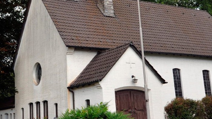 Weiße Kirche mit hohem Turm und braunem Dach, umgeben von Bäumen und Büschen