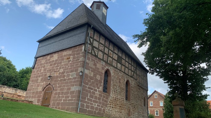 Kleines Steingebäude mit Rundbogenfenstern und einem steilen Dach, umgeben von Grünflächen.